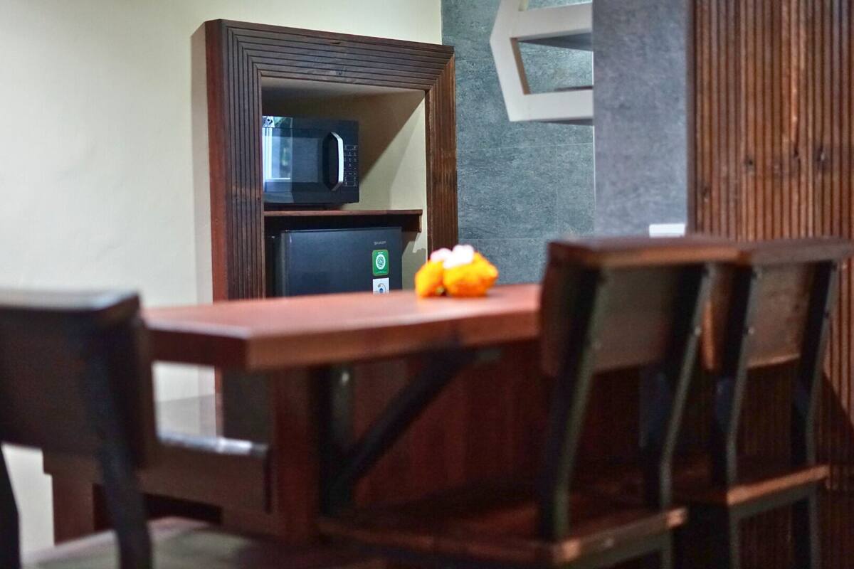 A dining area with a wooden table and chairs, a microwave above a refrigerator, and a small orange fruit on the table.