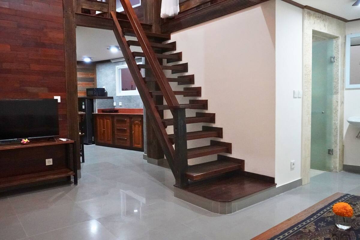 A living room with a wooden staircase, a TV, and a kitchen area in the background.