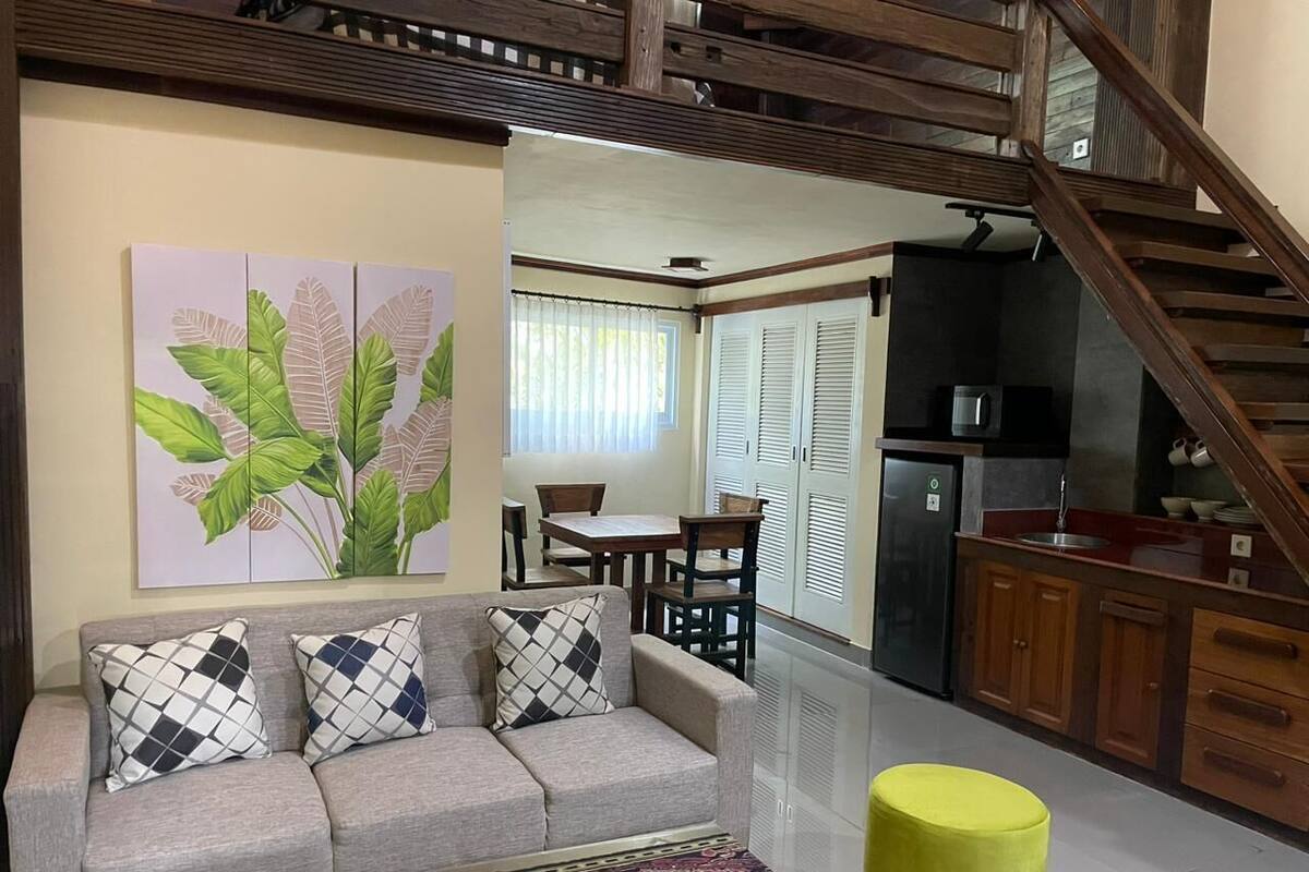 A living room with a sofa, patterned cushions, a coffee table, and a painting of leaves. A staircase leads to a kitchen area with wooden cabinets and a refrigerator.