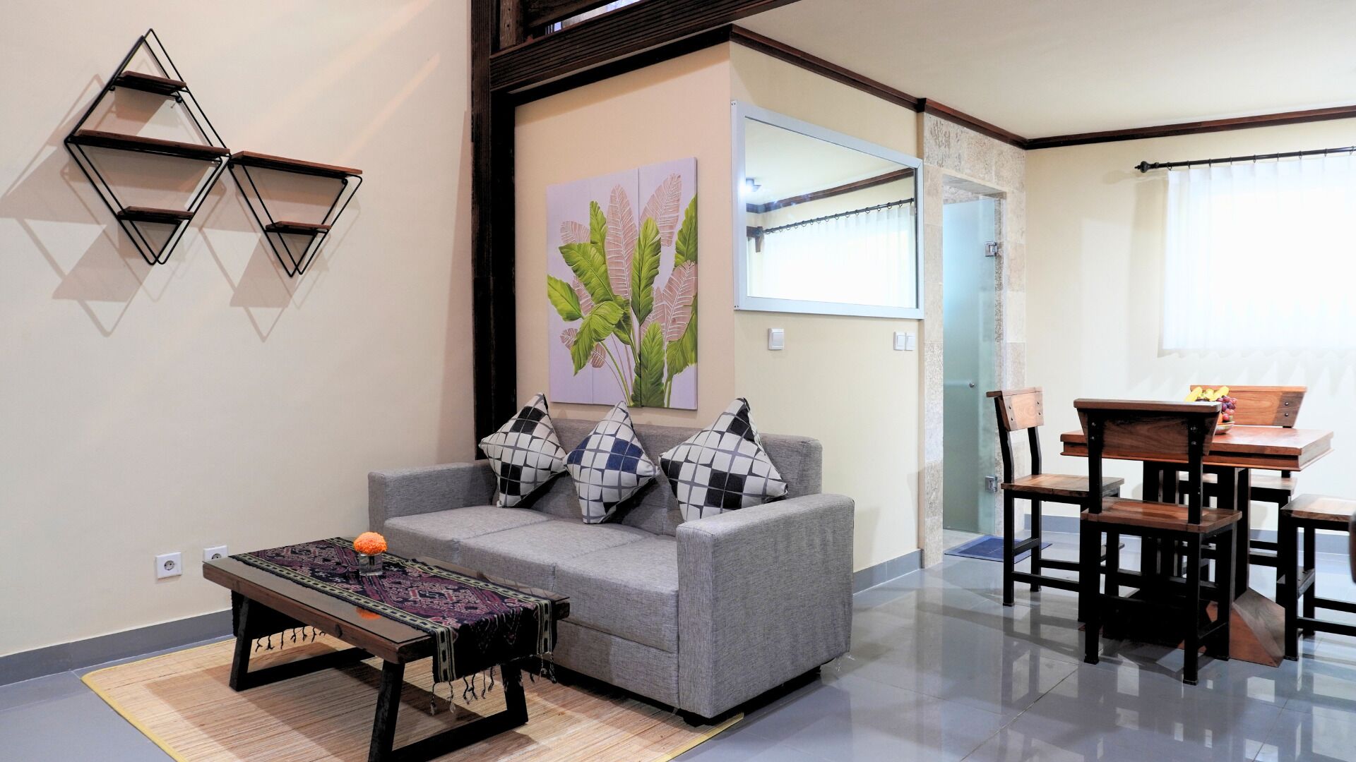 Uma sala de estar com um sofá cinza, almofadas estampadas, uma mesa de centro e um quadro de folhas na parede.