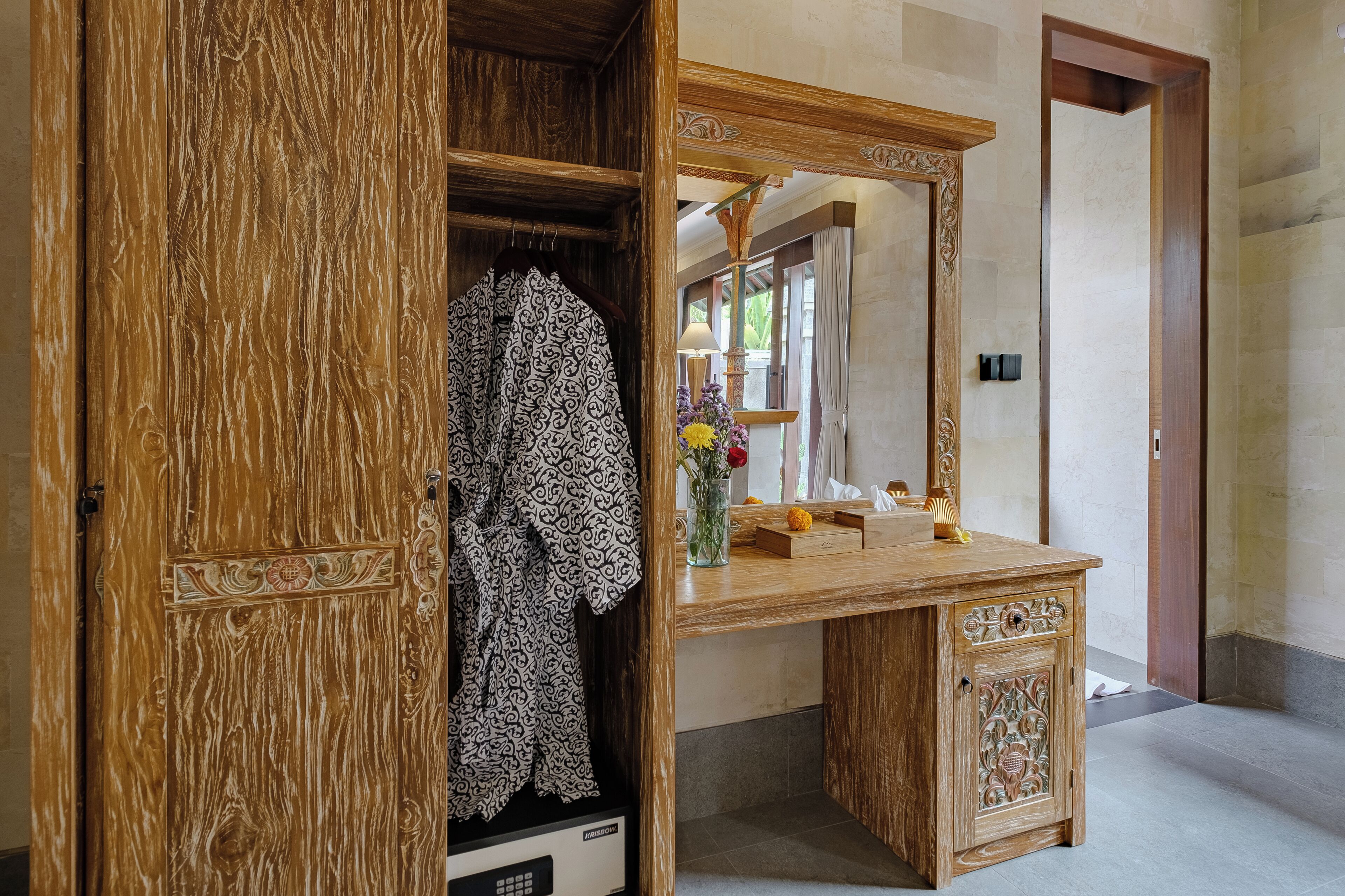 A wooden vanity with a mirror, a wooden wardrobe, and a vase with flowers.