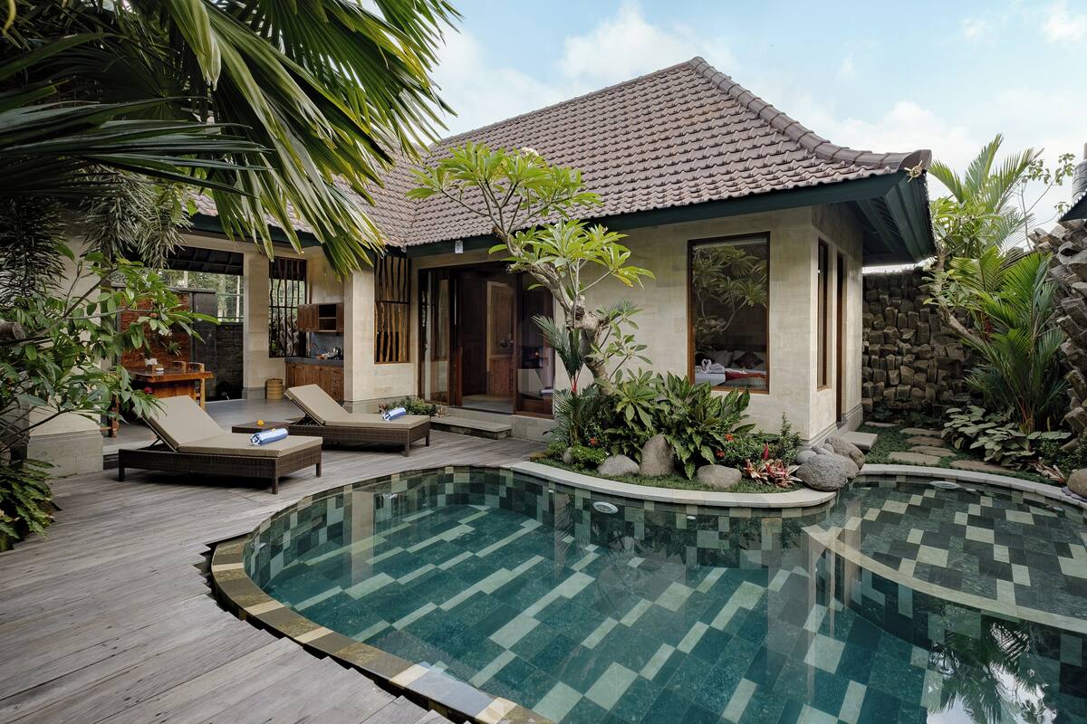 A pool area with lounge chairs, a small building with a tiled roof, and surrounding greenery.