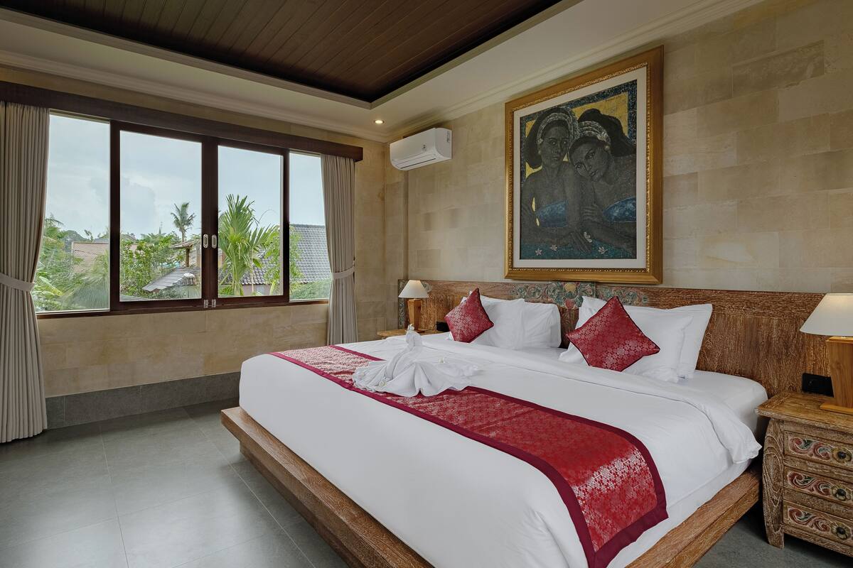 A hotel room with a large bed, a wooden headboard, a painting on the wall, and a view of greenery outside.