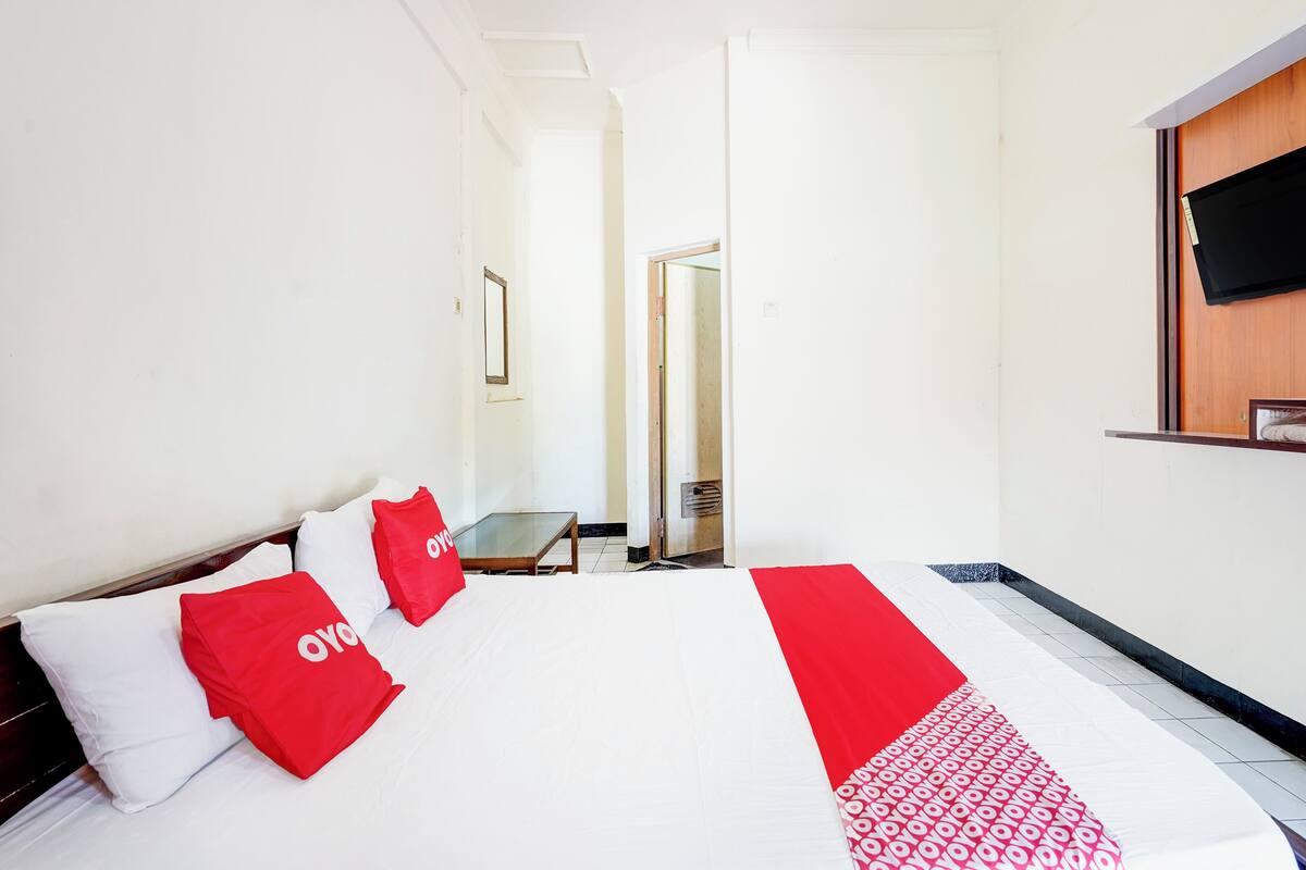 Hotelkamer met een bed, twee rode kussens, een televisie aan de muur en een deur naar een andere kamer.