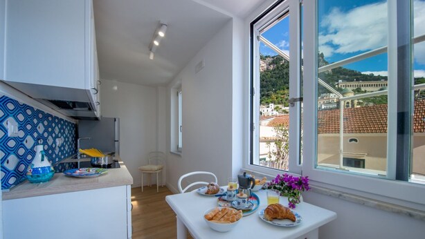 Cuina compacta amb armaris blancs, un frontissis amb disseny i una zona de dinar amb taula preparada per l'esmorzar. Una finestra ofereix vistes a un paisatge de muntanya.