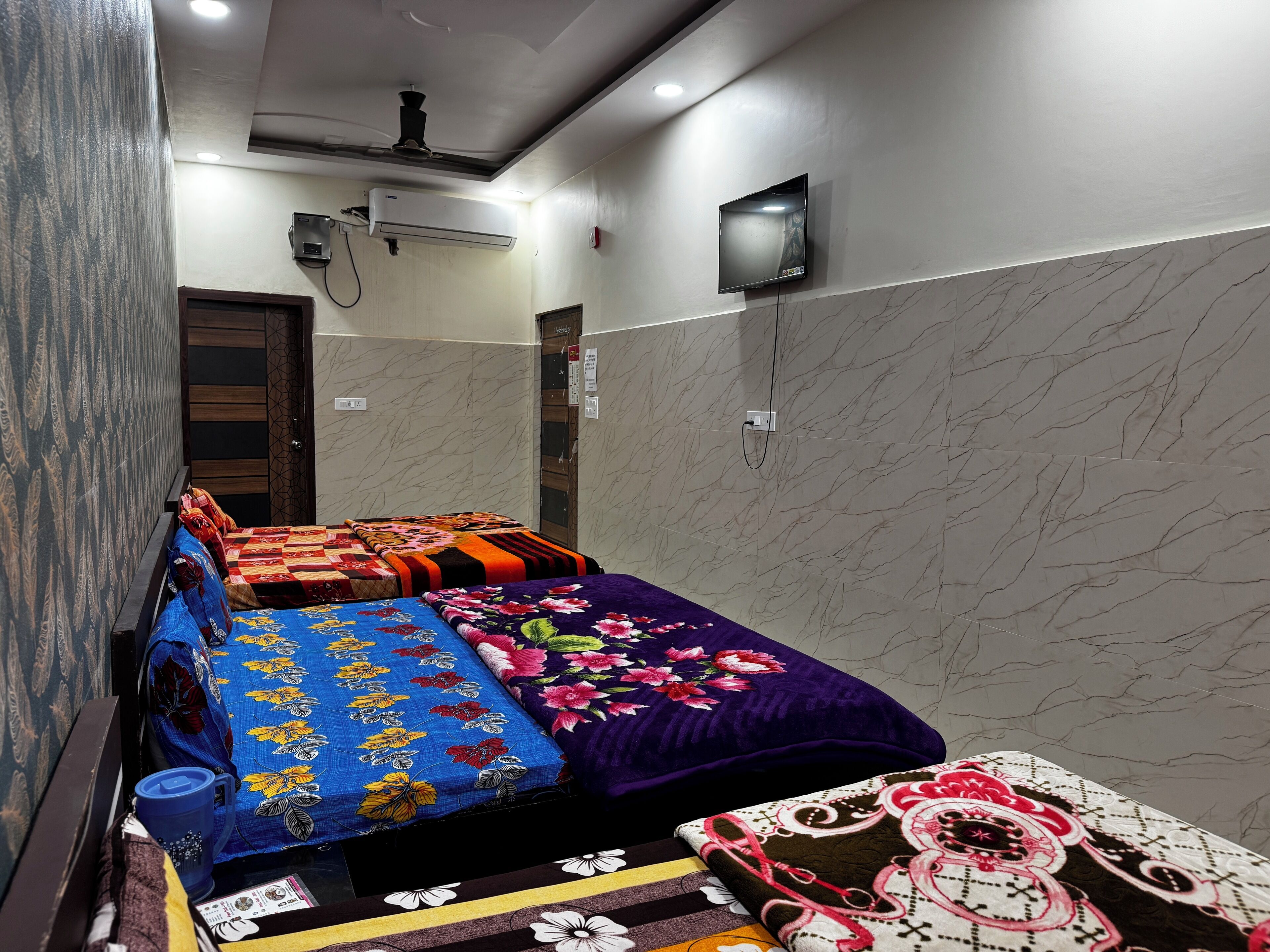 A bedroom with a bed covered in colorful floral bedding, a ceiling fan, an air conditioning unit, and a mounted TV.