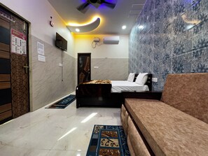 A hotel room with a bed, a sofa, a patterned rug, a ceiling fan, and a wall-mounted air conditioner. - Vrindavani Kunj (Vrindavan)