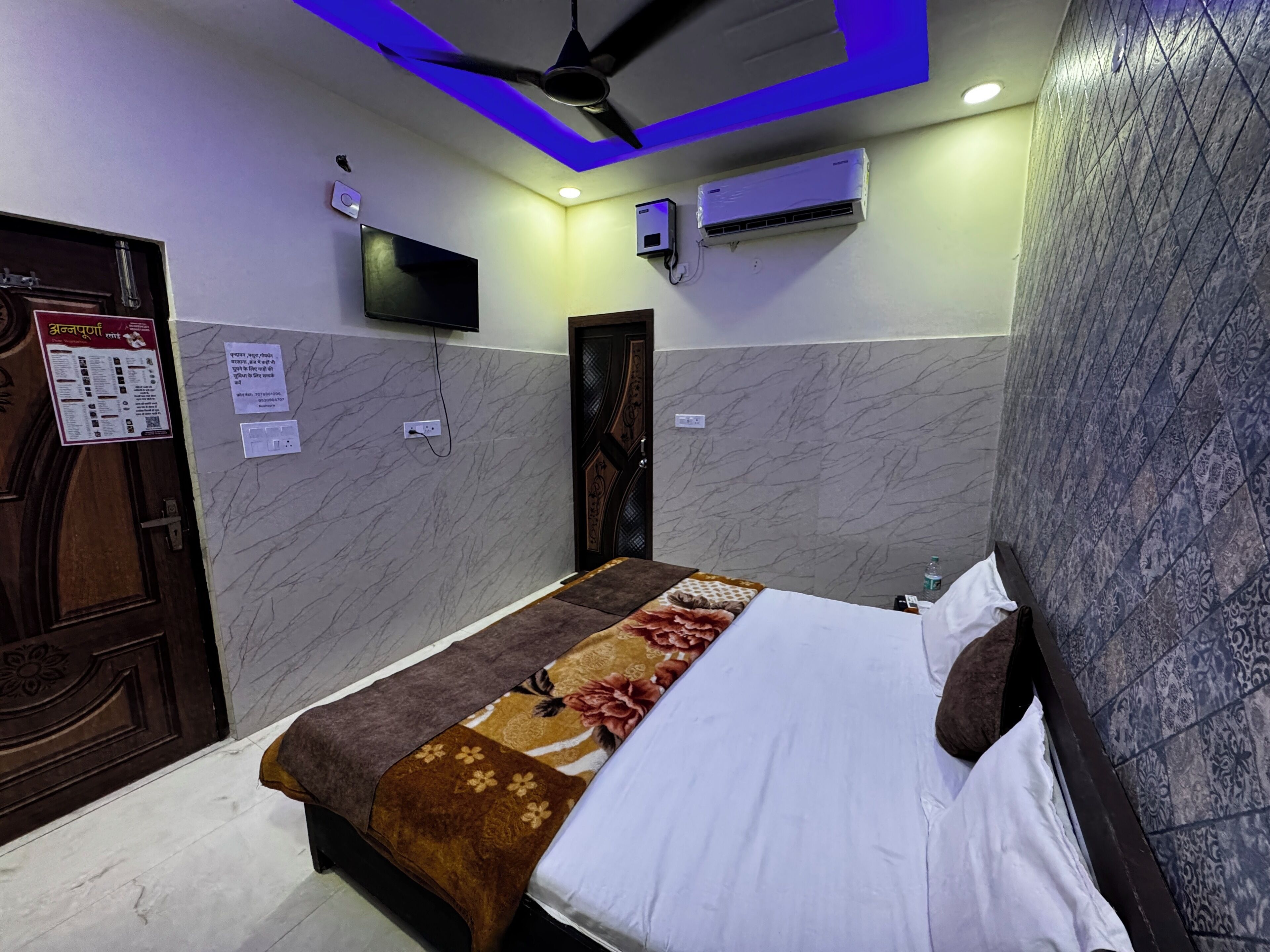 A hotel room with a bed, a ceiling fan, an air conditioning unit, a TV mounted on the wall, and a door with a wooden panel.