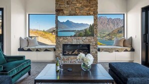 A living room with a stone fireplace, large windows with mountain views, a sofa, and a coffee table with a bottle and flowers.