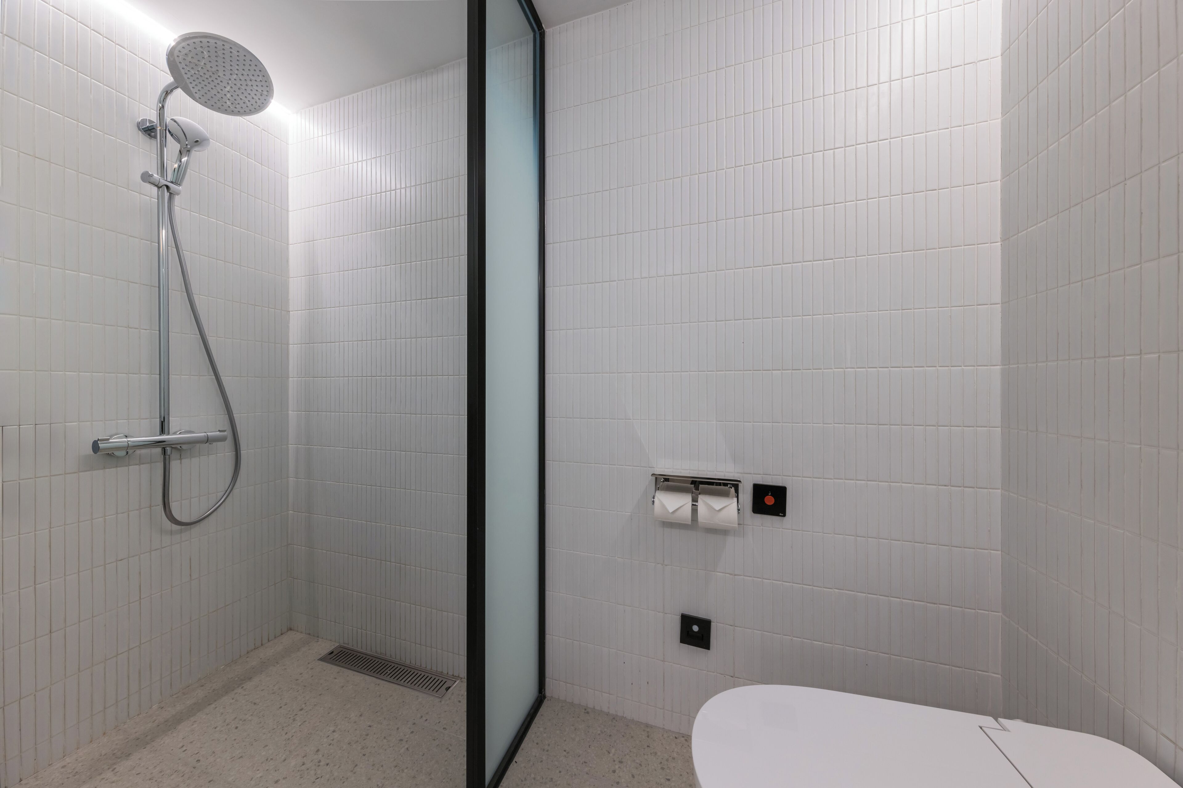 Un baño moderno con paredes de azulejos blancos, cabina de ducha con mampara de vidrio, inodoro y dispensador de toallas.