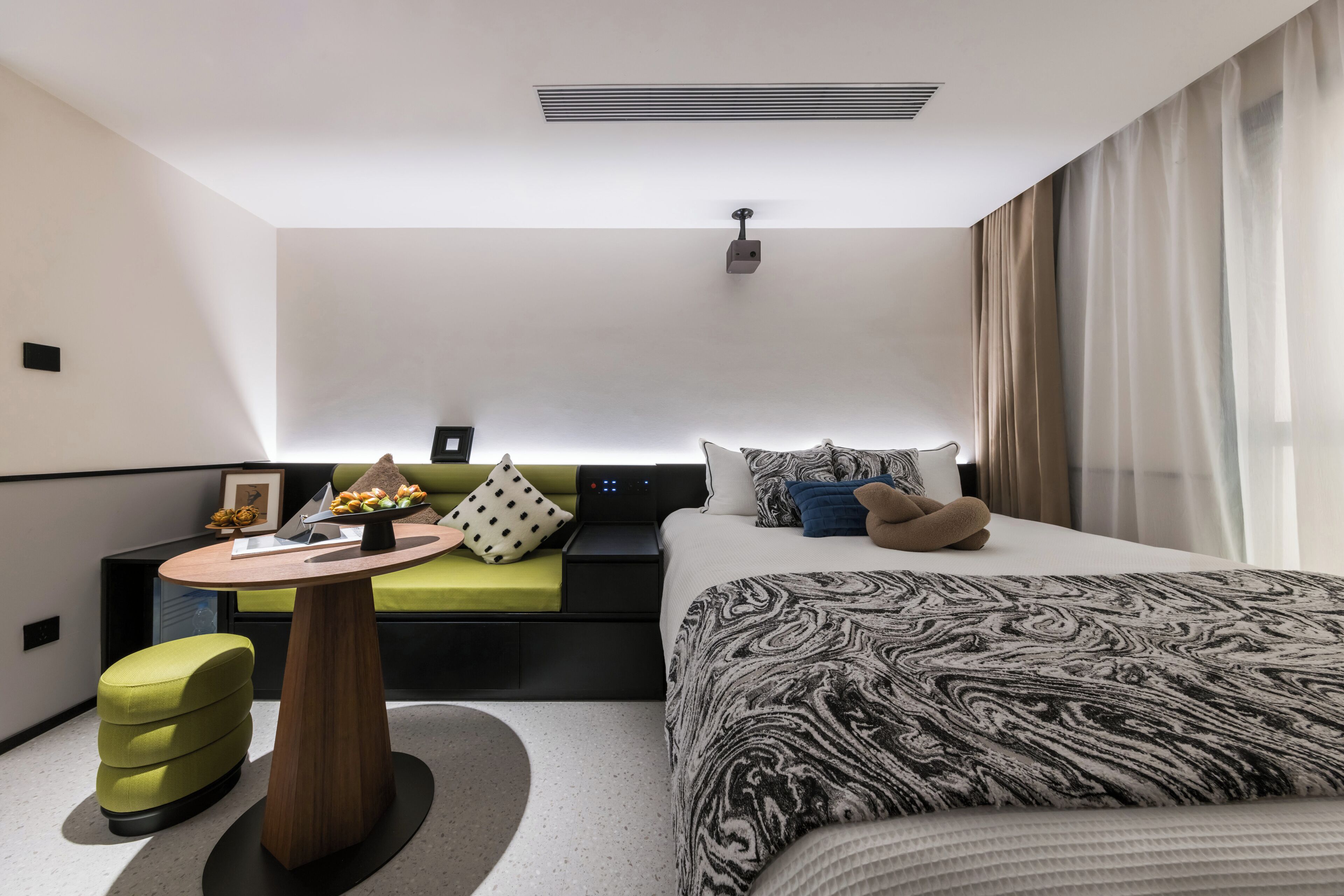 A modern hotel room with a bed, a sofa, a small table with a vase of flowers, and a projector on the ceiling.