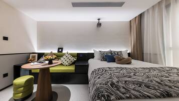 A modern hotel room with a bed, a sofa, a small table with a vase of flowers, and a projector on the ceiling.