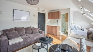 A modern living room with a grey sofa, a round coffee table, and a dining area with a small table and chairs. There is a kitchen area with wooden cabinets and a window. - New Apartment Center of Paris/le Marais (Paris)