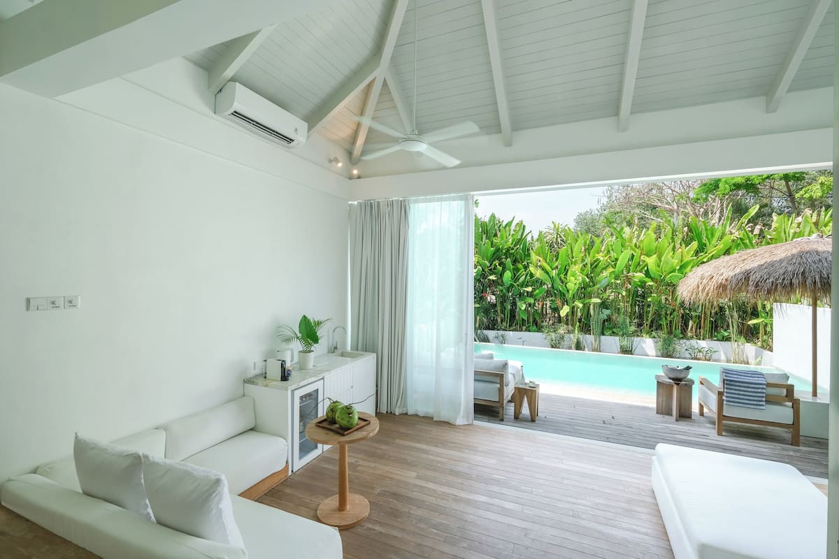 Una stanza moderna e luminosa, con un divano bianco, un tavolino con una pianta e vista su una piscina e vegetazione lussureggiante.