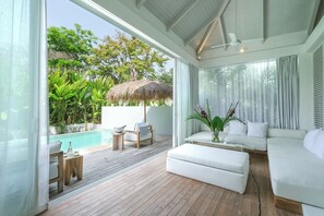A modern living room with a wooden floor, white sofas, a small table, and a view of a swimming pool and lush greenery. - Sacred and Temple Bungalow (Canggu)