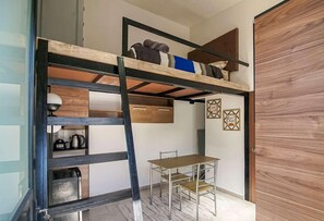 A room with a bunk bed, a desk, and a chair. There is a window, a door, and a wall with two framed pictures.