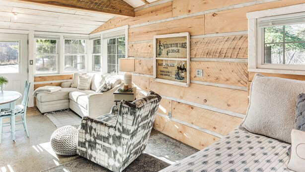Een gezellig interieur van een blokhut met een bank, fauteuil en een bordje met de tekst 'Welcome to the Lake Lodge'.