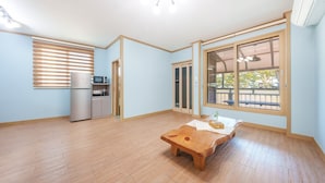 A spacious room with a wooden floor, a built-in kitchen with a refrigerator and microwave, a large window with a view of the outdoors, and a wooden table with a plant. - Gyeongju Beach Castle Pension (Gyeongju)