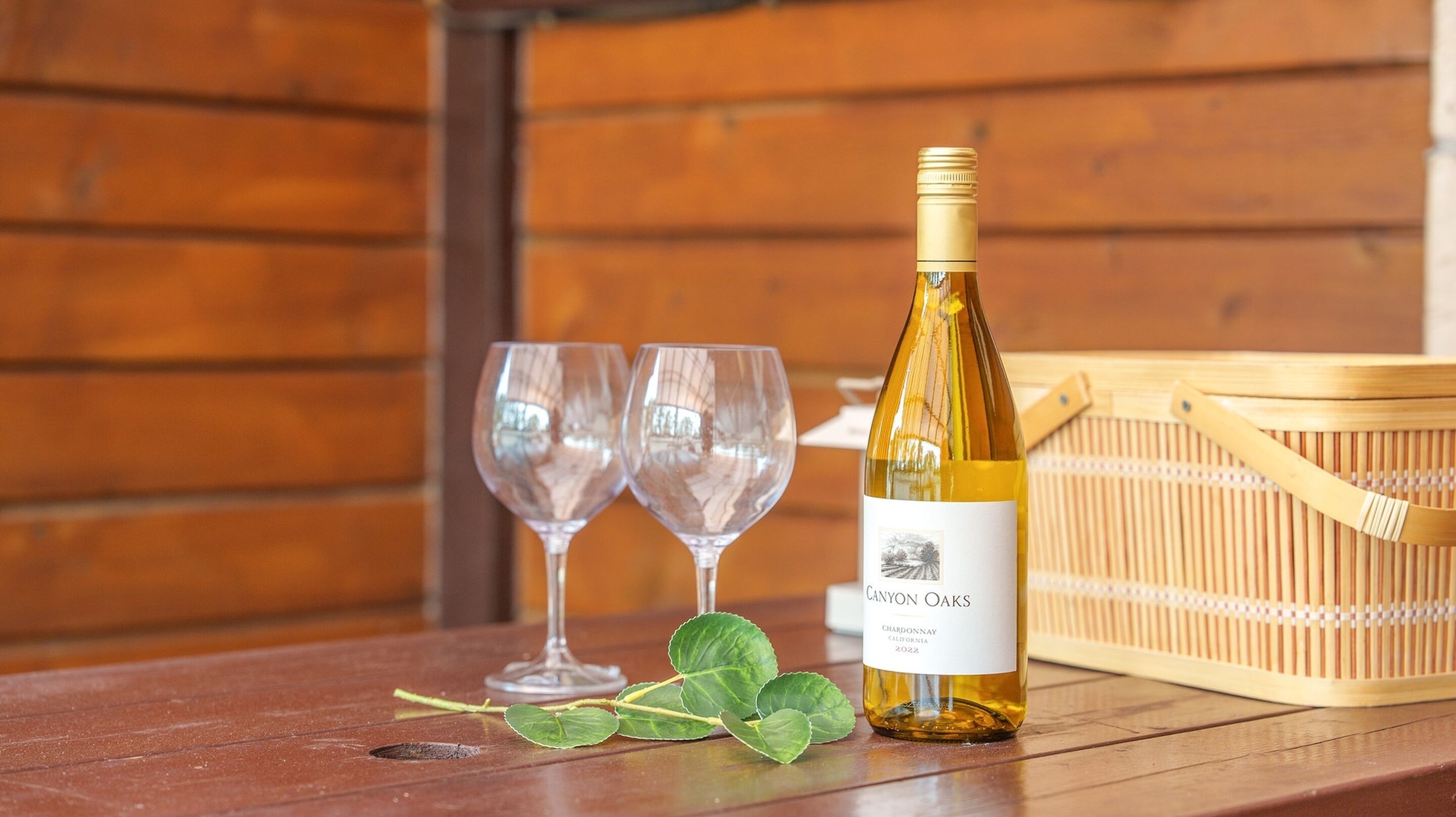 A bottle of Canyon Oaks Chardonnay 2022 wine, two empty wine glasses, and a wooden basket on a wooden table.
