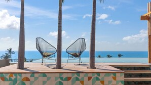 Ein Poolbereich mit zwei Stühlen aus Drahtgestell, die auf den Ozean ausgerichtet sind, umgeben von Palmen und einer gefliesten Terrasse.