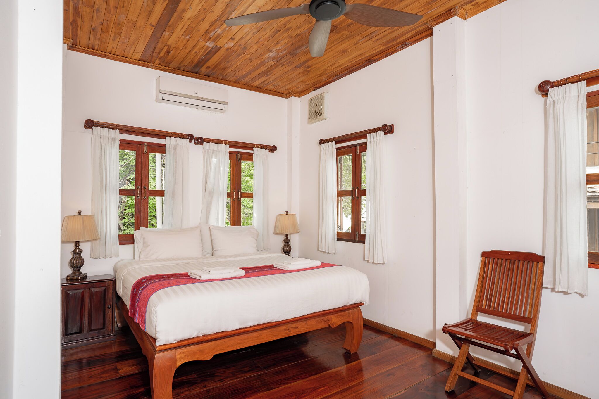 A bedroom with a wooden bed, two windows with curtains, a ceiling fan, and a wooden chair.