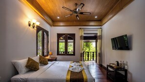 Une chambre d’hôtel avec un plafond en bois, un lit recouvert de draps blancs et doté d’une tête de lit décorative, un téléviseur fixé au mur et un balcon offrant une vue sur la verdure.