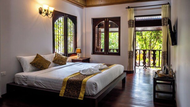 Un lit bien fait, avec une couvre-lit à motifs et des oreillers, une lampe de chevet, et une vue sur un balcon avec une balustrade et de la verdure à l’extérieur.