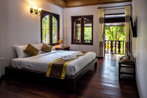 A neatly made bed with a patterned bedspread and pillows, a bedside lamp, and a view of a balcony with a railing and greenery outside. - Villa Mira Heritage  (Luang Prabang)