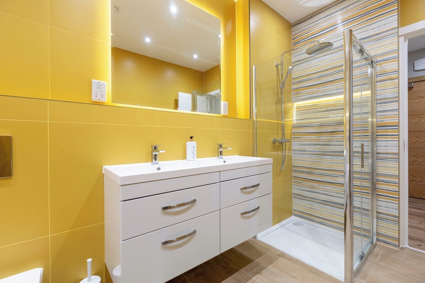 Une salle de bain moderne avec des murs jaunes, un meuble-lavabo blanc doté de deux vasques et une cabine de douche en verre.