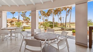 Un espacio de comedor al aire libre cubierto, con sillas blancas y una mesa redonda, con vistas a un jardín ajardinado con palmeras y una edificación con tejado de tejas.