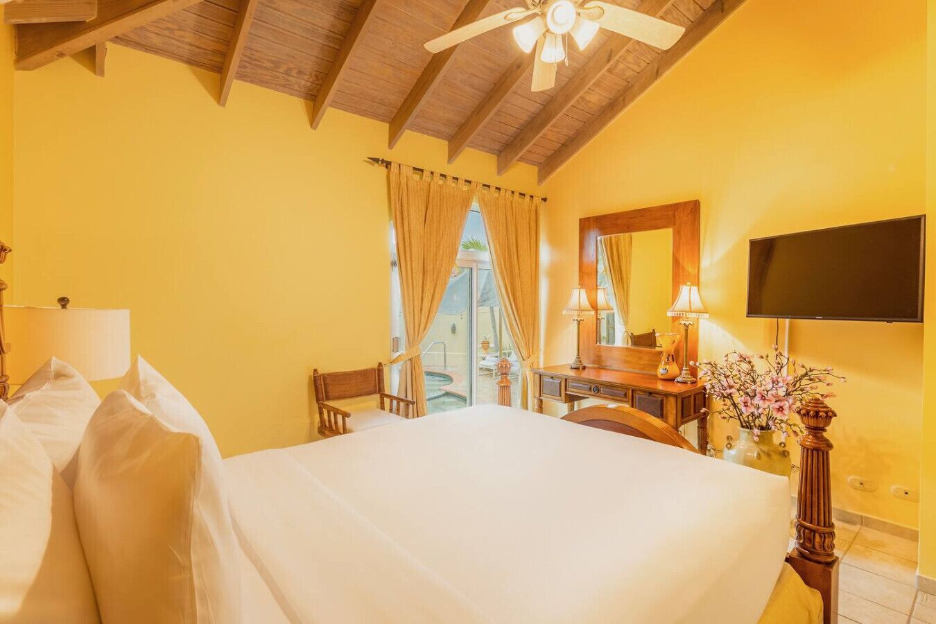 A hotel room with a large bed, a wooden ceiling, a desk with a chair, a TV mounted on the wall, and a window with curtains.