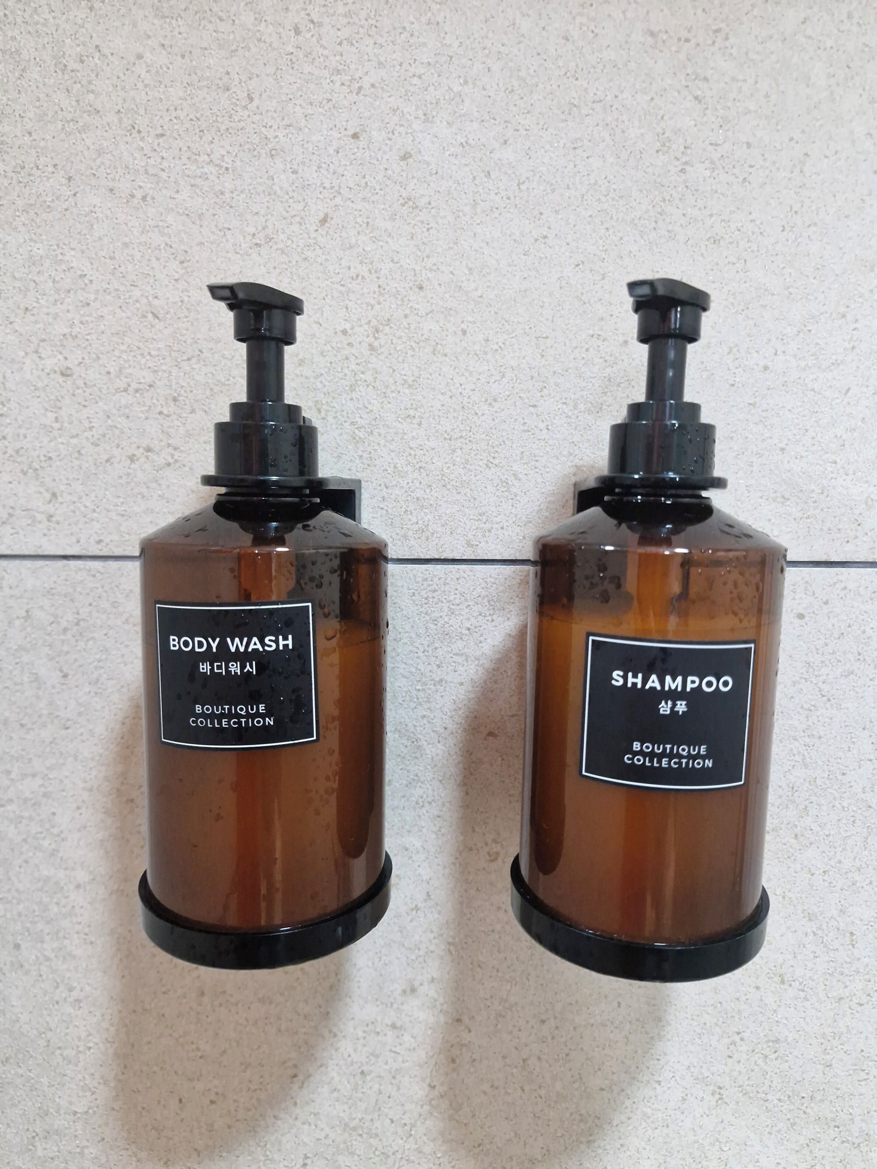 To flasker bodywash og shampoo med sorte pumper, mærket både på engelsk og koreansk, foran en struktureret væg.