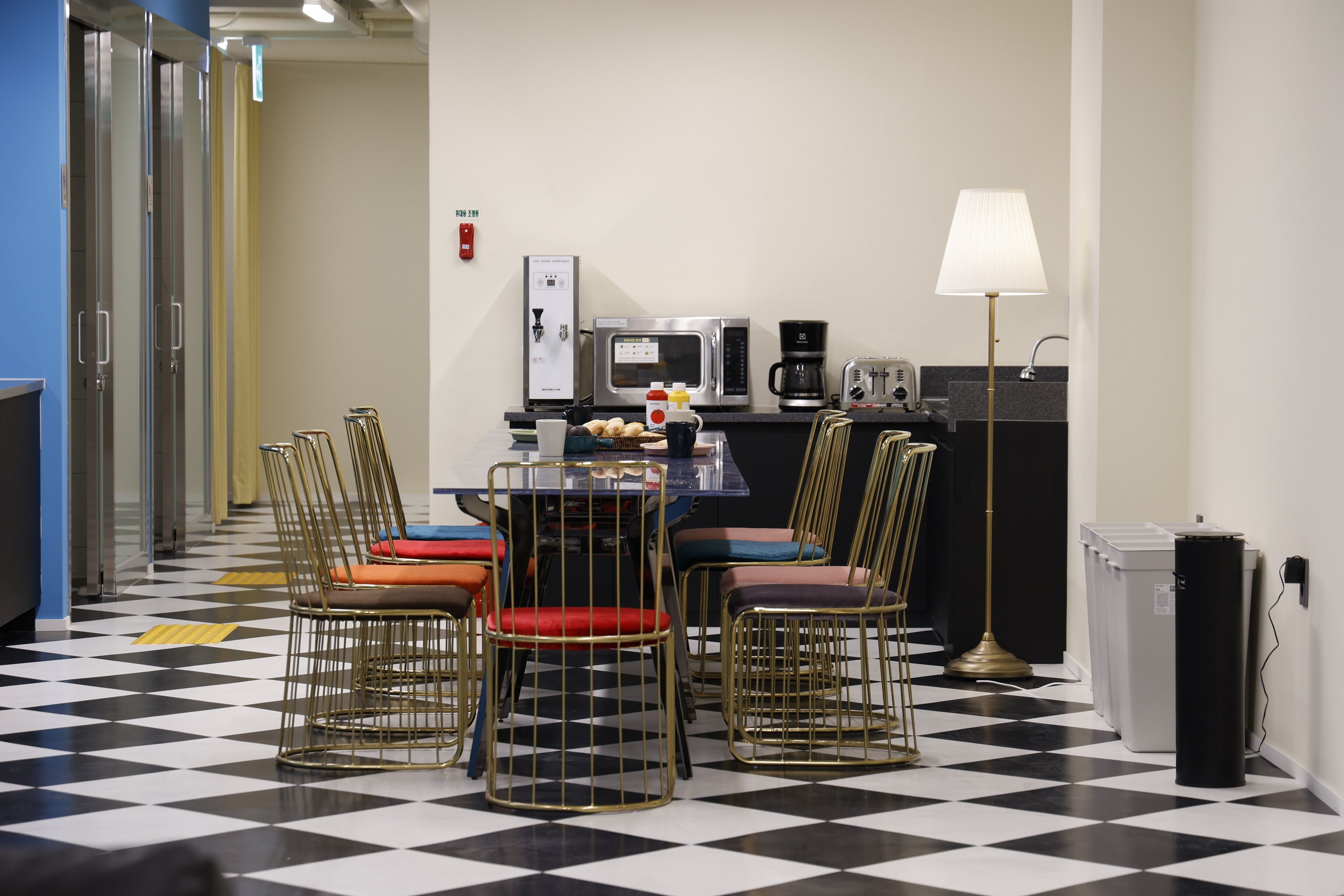 Une chambre d’hôtel avec un sol à carreaux, une table à manger avec des chaises, un four à micro-ondes, une machine à café, une lampe et une poubelle.
