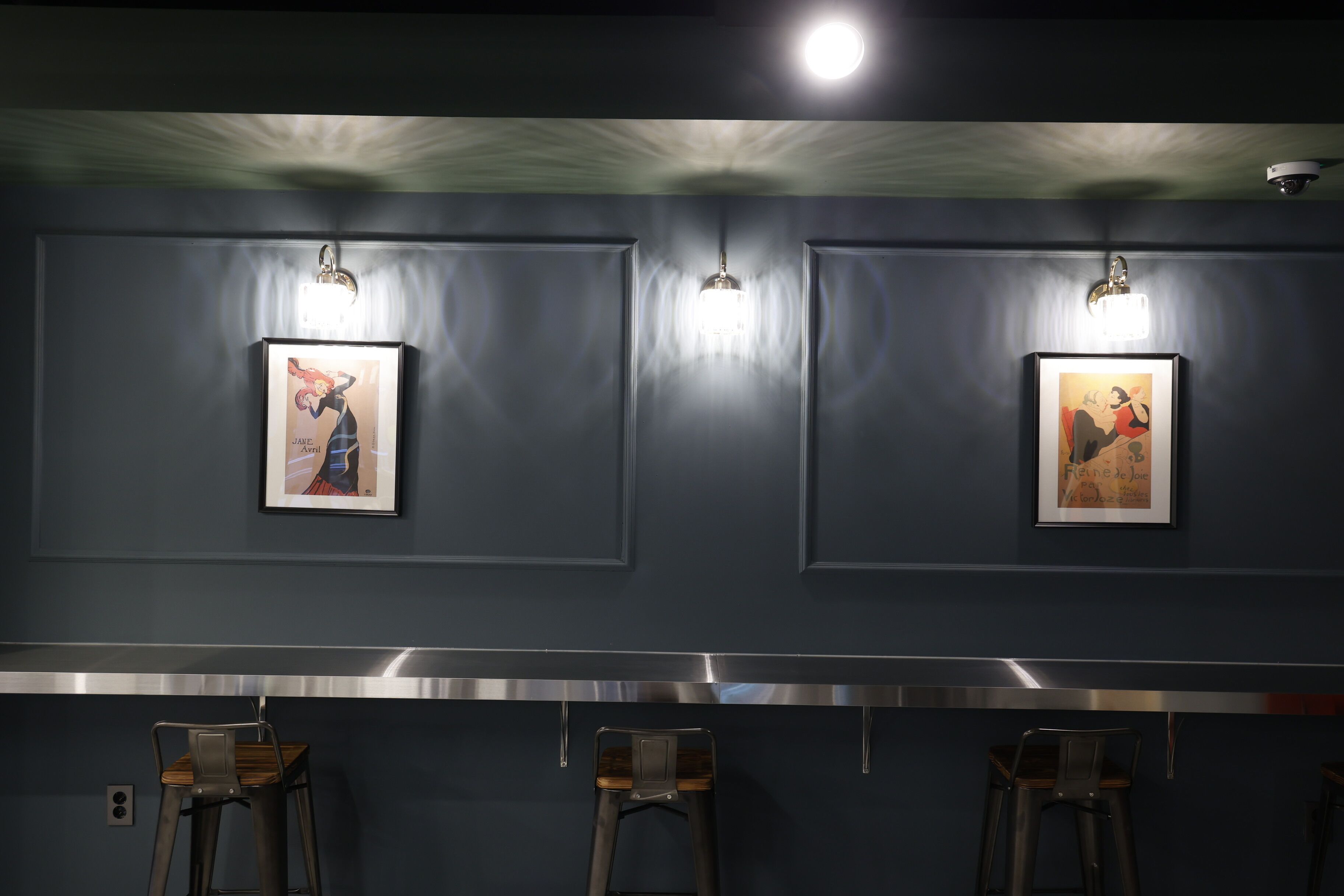 Un área de bar con dos cuadros en la pared, una barra de acero inoxidable y tres taburetes.