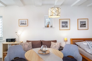 Um quarto aconchegante com uma mesa de madeira redonda, um sofá e uma cama. Há quadros nas paredes, uma janela e uma luminária em uma mesinha de apoio.