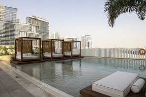 Una piscina en la azotea con tres cabañas de madera y un área de descanso con un colchón y almohadas.