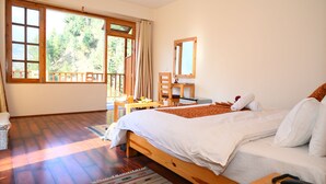 A bedroom with a large bed, wooden flooring, a desk with a chair, a window with a view of trees, and a mirror on the wall.