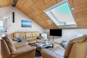 A cozy living room with a high ceiling, a large window, a flat-screen TV, and a leather sofa.