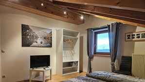 Ein Schlafzimmer mit Bett, Fernseher, Holzboden und einem Fenster mit Vorhängen, sowie einem Gemälde mit einer Berglandschaft.