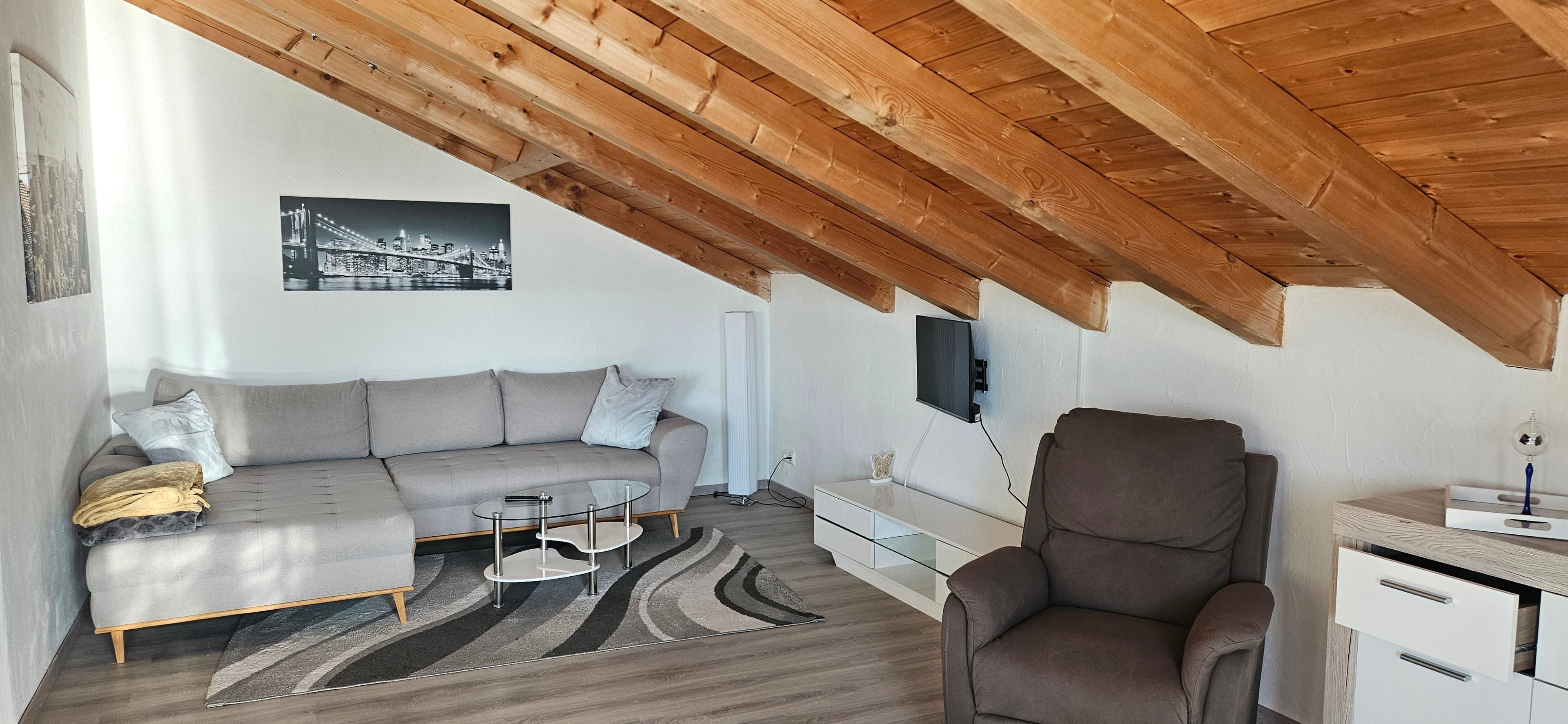 A modern loft-style living space with a high ceiling, a sofa, a coffee table, a chair, and a flat-screen TV mounted on the wall.