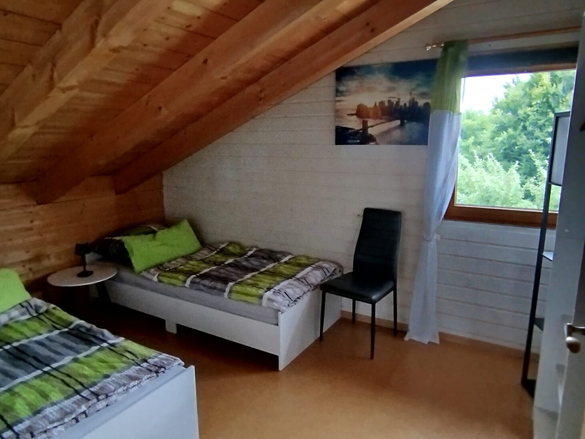 Een slaapkamer met een schuine kap, een bed met een groene en witte spreideken, een stoel, een klein rond tafeltje en een raam met uitzicht op groen.