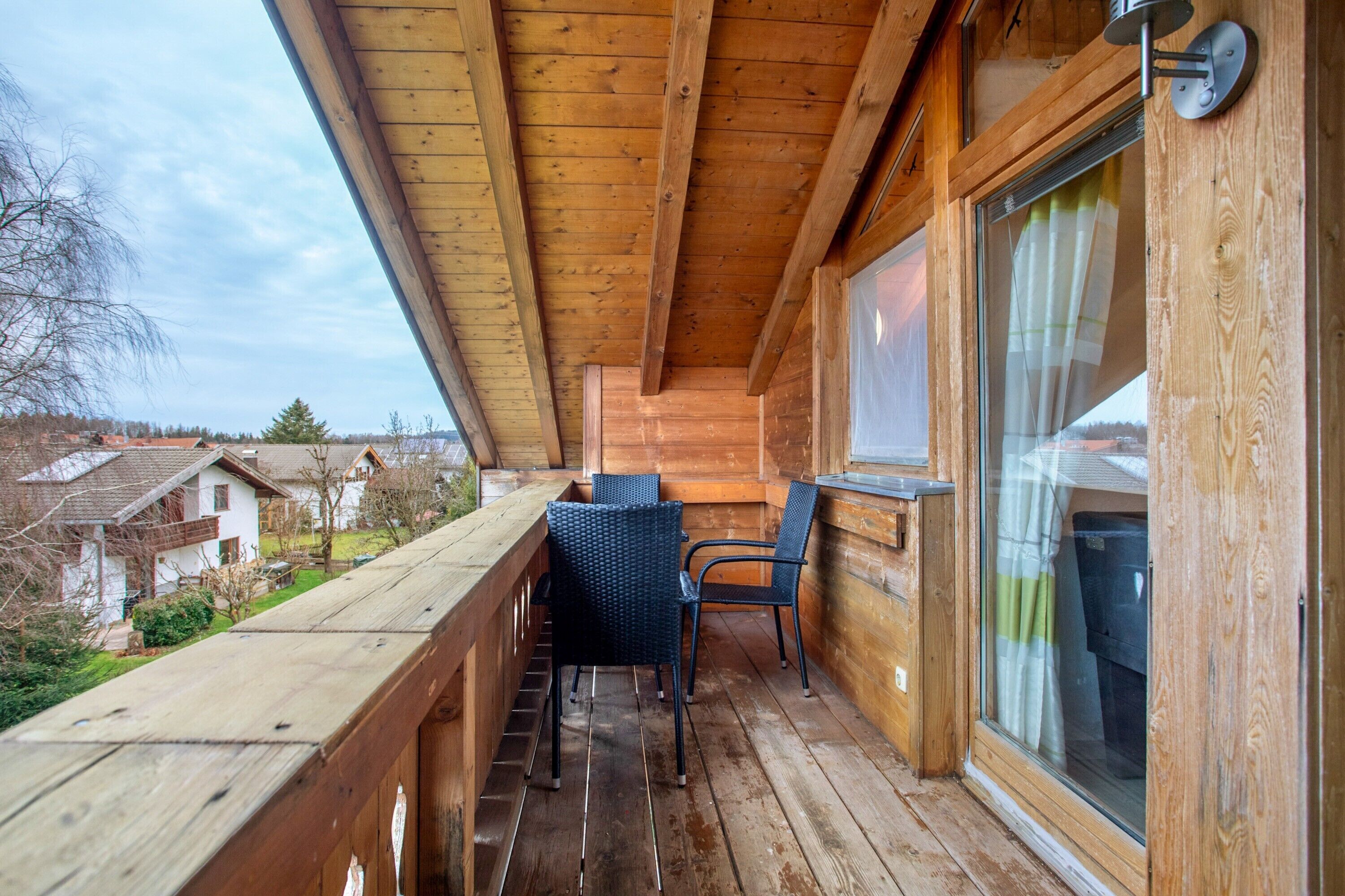 A wooden balcony with a table and chairs, a window with curtains, and a view of houses in the background.