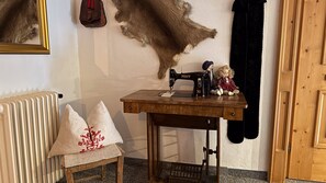 Una máquina de coser sobre una mesa de madera, un radiador, una silla con cojín, y una pared decorada con un abrigo de piel y una muñeca.
