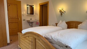 A bedroom with two wooden beds, a wooden wardrobe, a bathroom with a sink and mirror, and a wall-mounted light fixture.