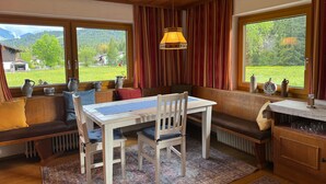 A cozy room with a wooden ceiling, a dining table with chairs, a bench with cushions, and large windows offering a view of a green landscape.