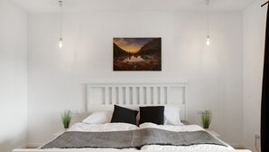 Een slaapkamer met een witte hoofdbord, een bed met wit en zwart beddengoed, twee kamerplanten en een schilderij van een landschap boven het bed.