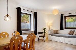 A room with a wooden dining table, chairs, a sofa, a potted plant, and a window with curtains.