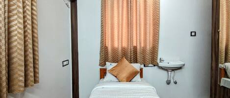 A hotel room with a bed, a bedside table, a window with curtains, a wall-mounted air conditioning unit, and a sink.
