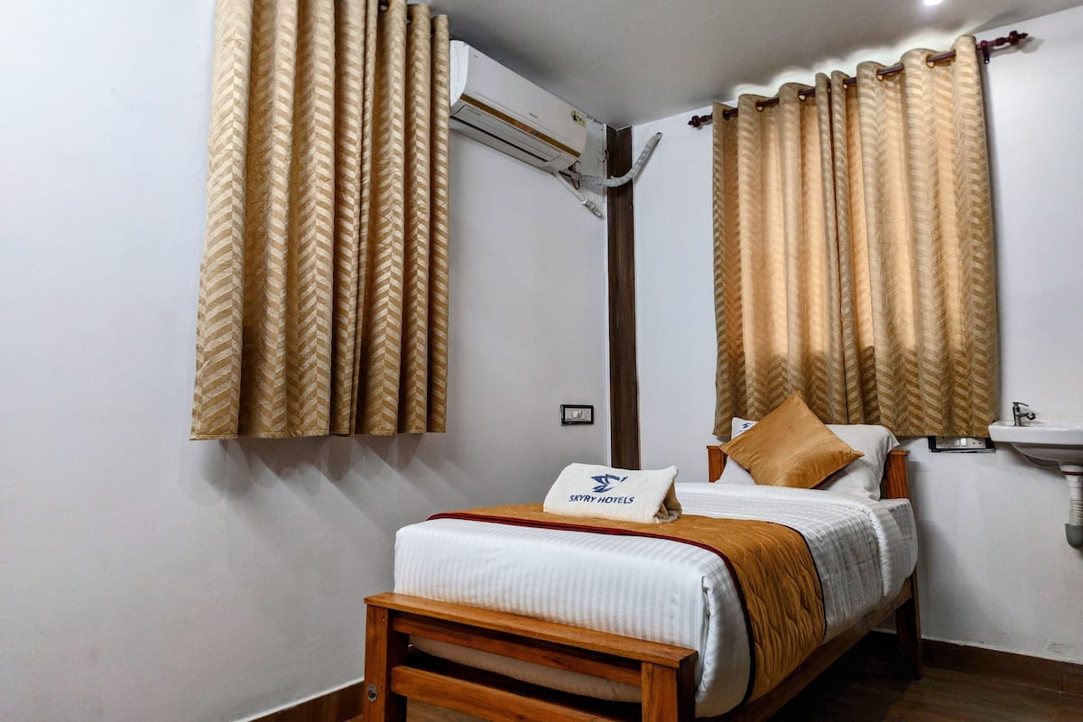 Une chambre d’hôtel comprenant un lit, une tête de lit en bois, une table de chevet, une petite table, un climatiseur fixé au mur, une salle de bain avec un lavabo et un miroir, et des rideaux à motifs chevrons.