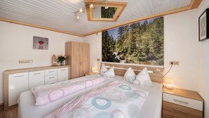 A bedroom with a bed, wooden headboard, bedside tables, a window with a forest view, and a painting on the wall. - Apartment 'Steinbühlschlössle' with Shared Terrace, Shared Garden and Wi-Fi (Schönwald im Schwarzwald)